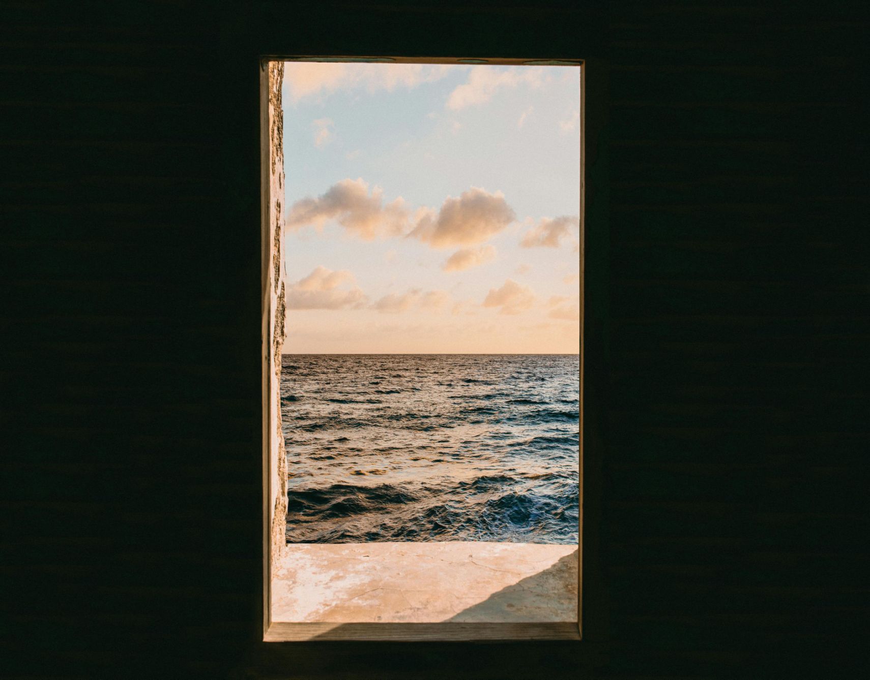 Fenêtre rectangulaire s'ouvrant sur un paysage marin calme et ensoleillé depuis une pièce sombre